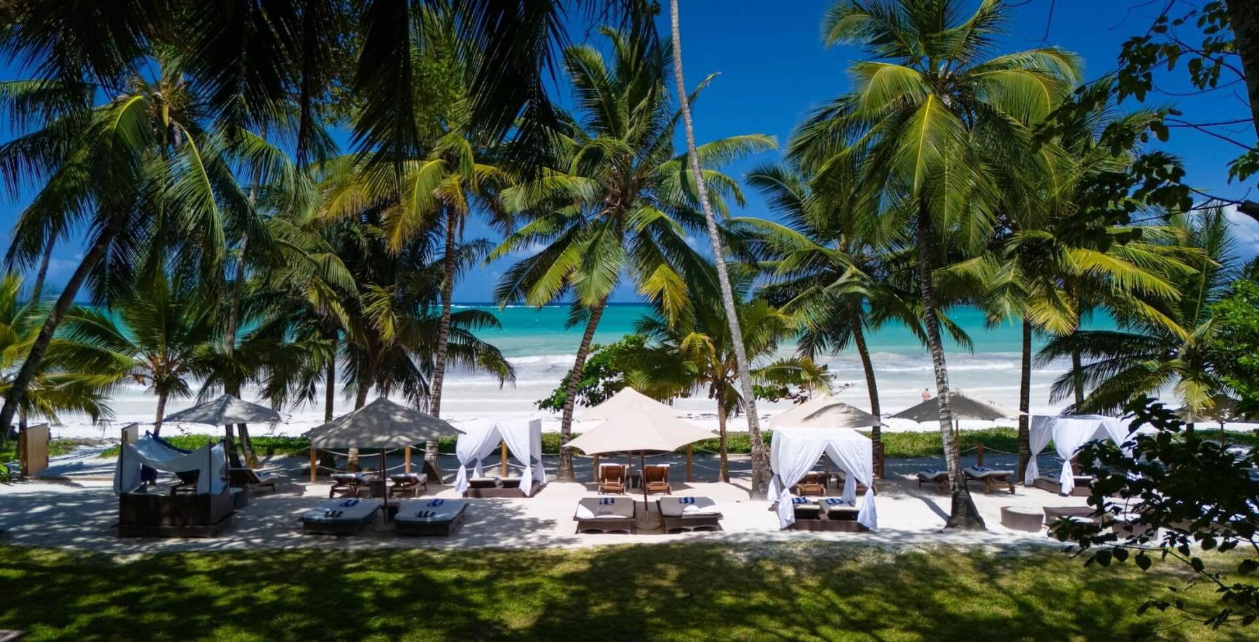 Shaded day beds and loungers beneath palm trees on the white sand at Almanara, with the turquoise Indian Ocean beyond