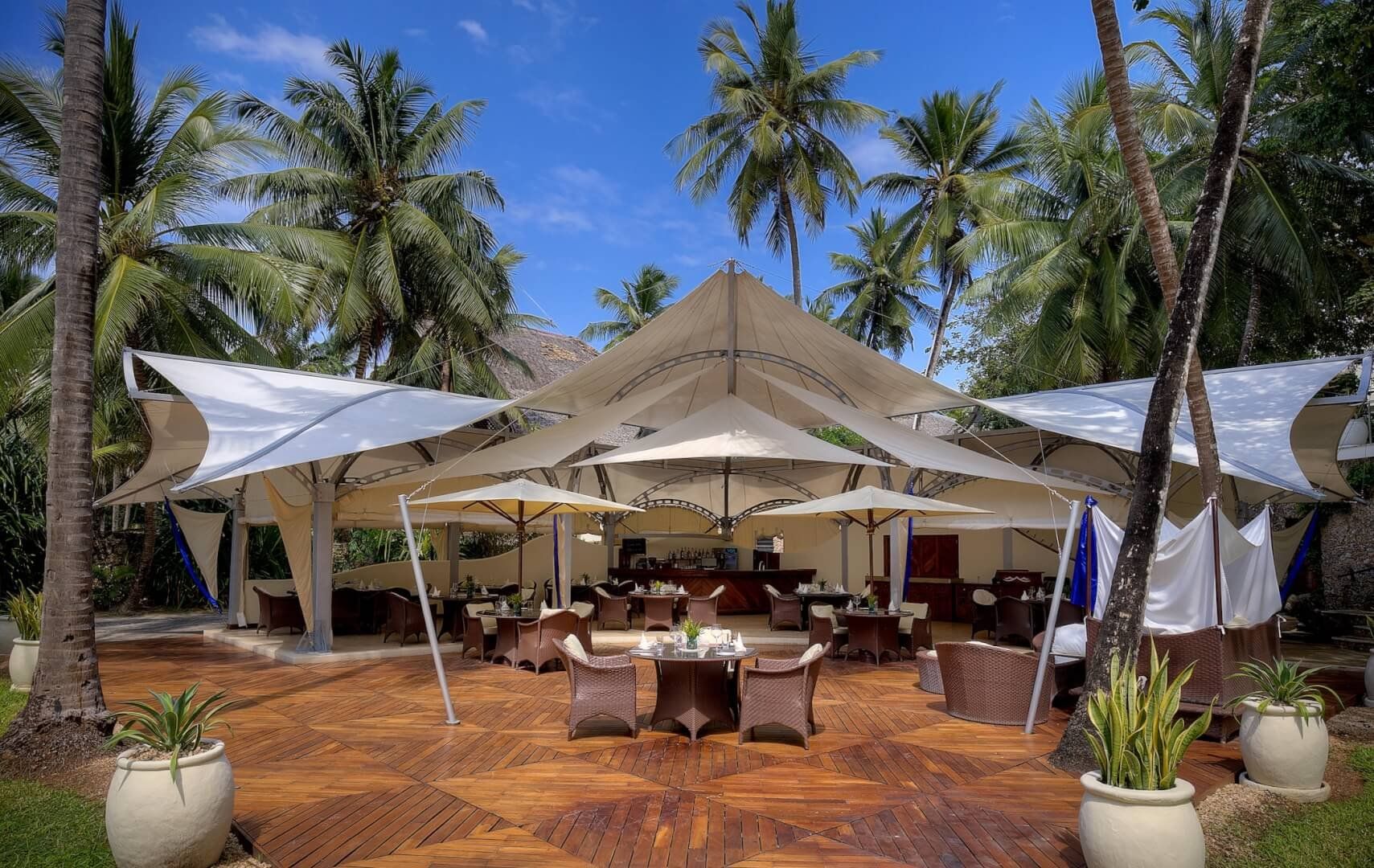 Sails, Almanara’s beach restaurant, with tables on a wooden deck under white canopies and palms
