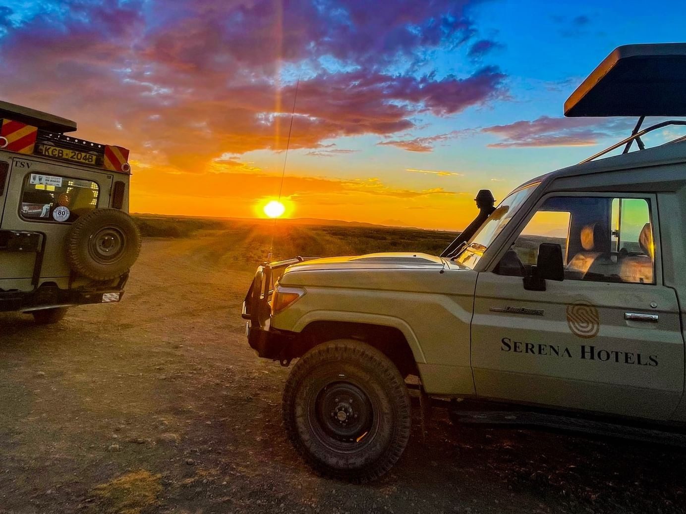 Serena game drive vehicles parked on a dirt track at sunset, with the sun low on the horizon