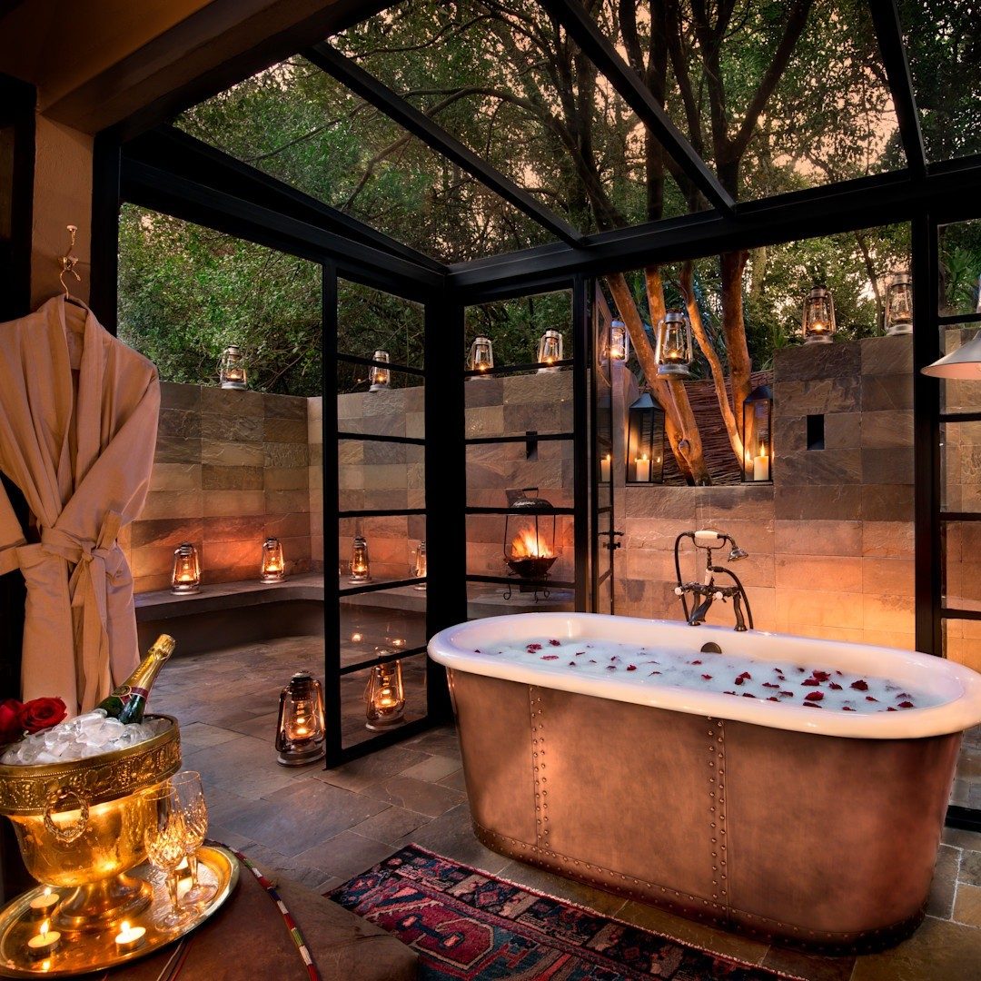 Copper bathtub in a glass-walled bathroom lit by lanterns at dusk at &Beyond Bateleur Camp, with an ice bucket beside it
