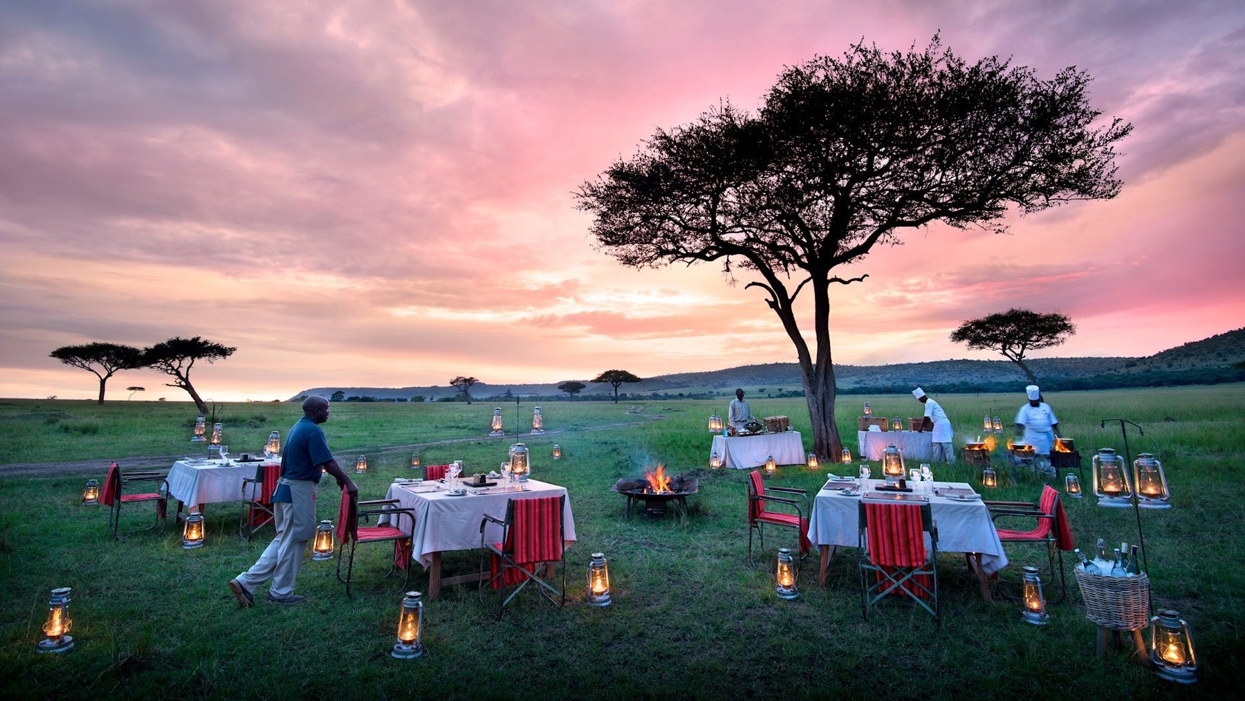 Dinner tables with red chairs and lanterns set on the grass under an acacia at sunset near &Beyond Bateleur Camp