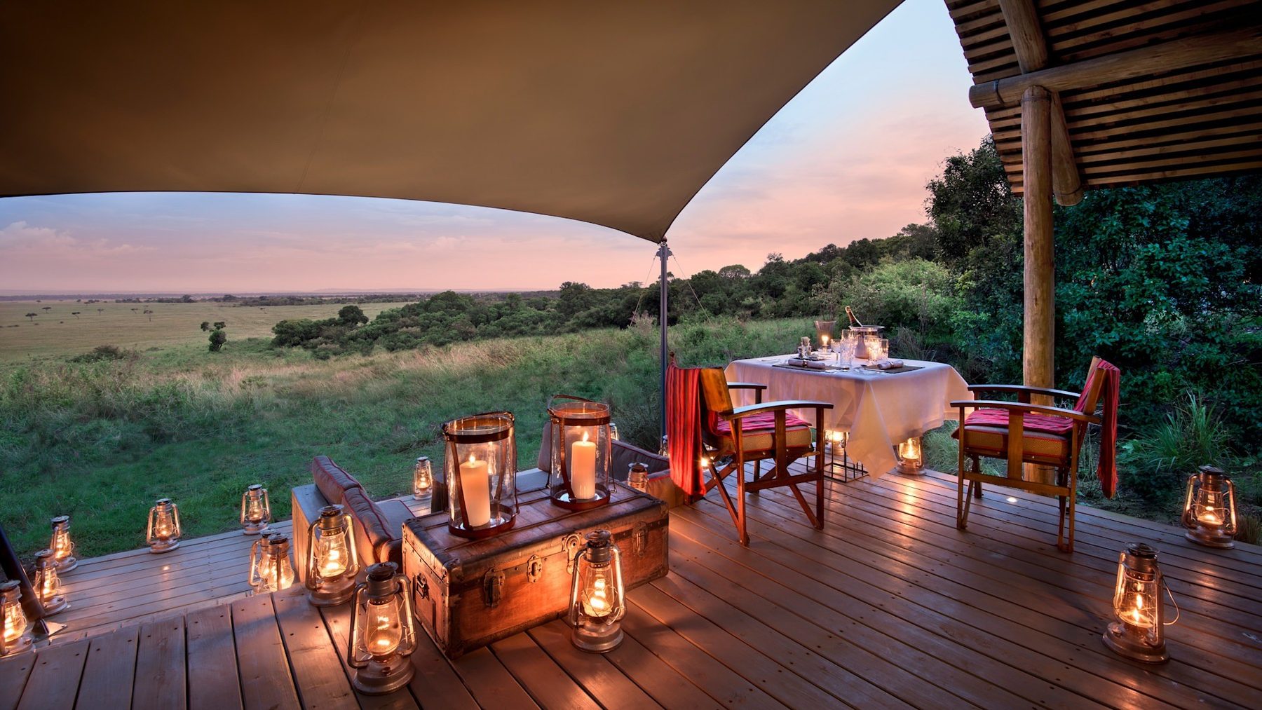 Tables set with lanterns on a wooden deck under a canvas shade at &Beyond Bateleur Camp, overlooking green plains