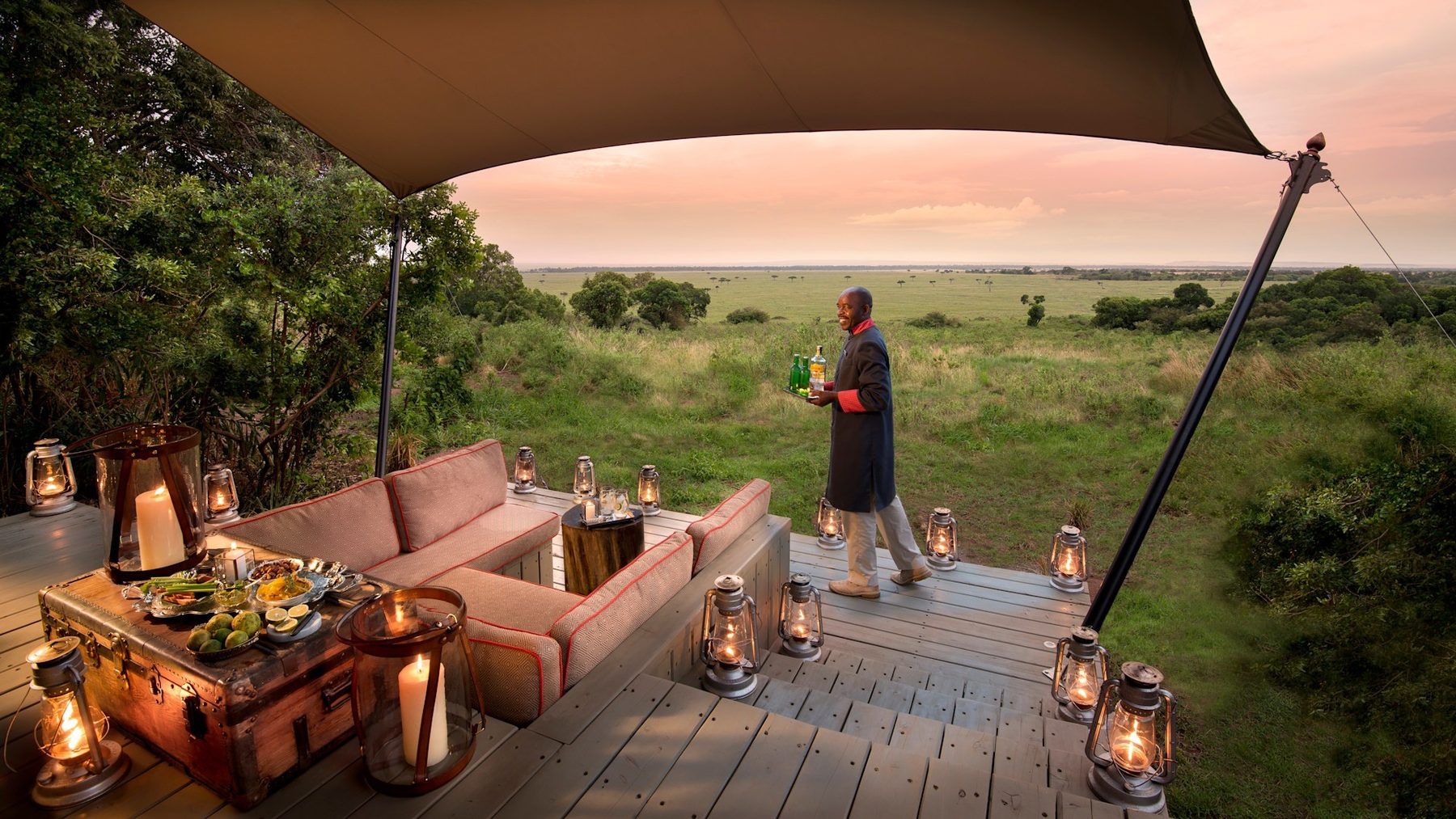 Sofas and lanterns on a shaded deck at &Beyond Bateleur Camp, with a waiter carrying drinks and plains stretching beyond