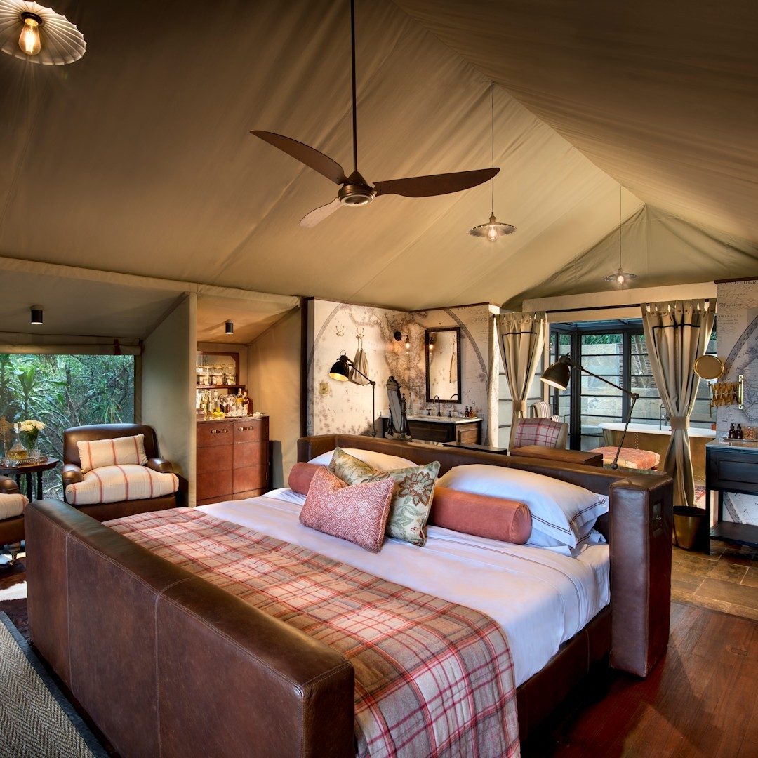Tented suite at &Beyond Bateleur Camp with a leather bed, plaid throw, ceiling fan and a writing desk by the window