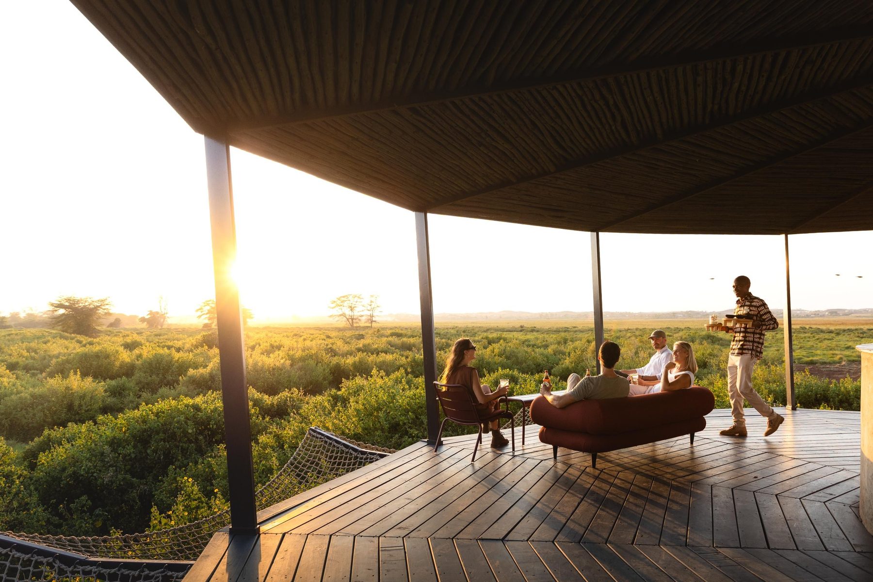 Guests relaxing on a shaded wooden deck at Angama Amboseli as the sun sets over green bush