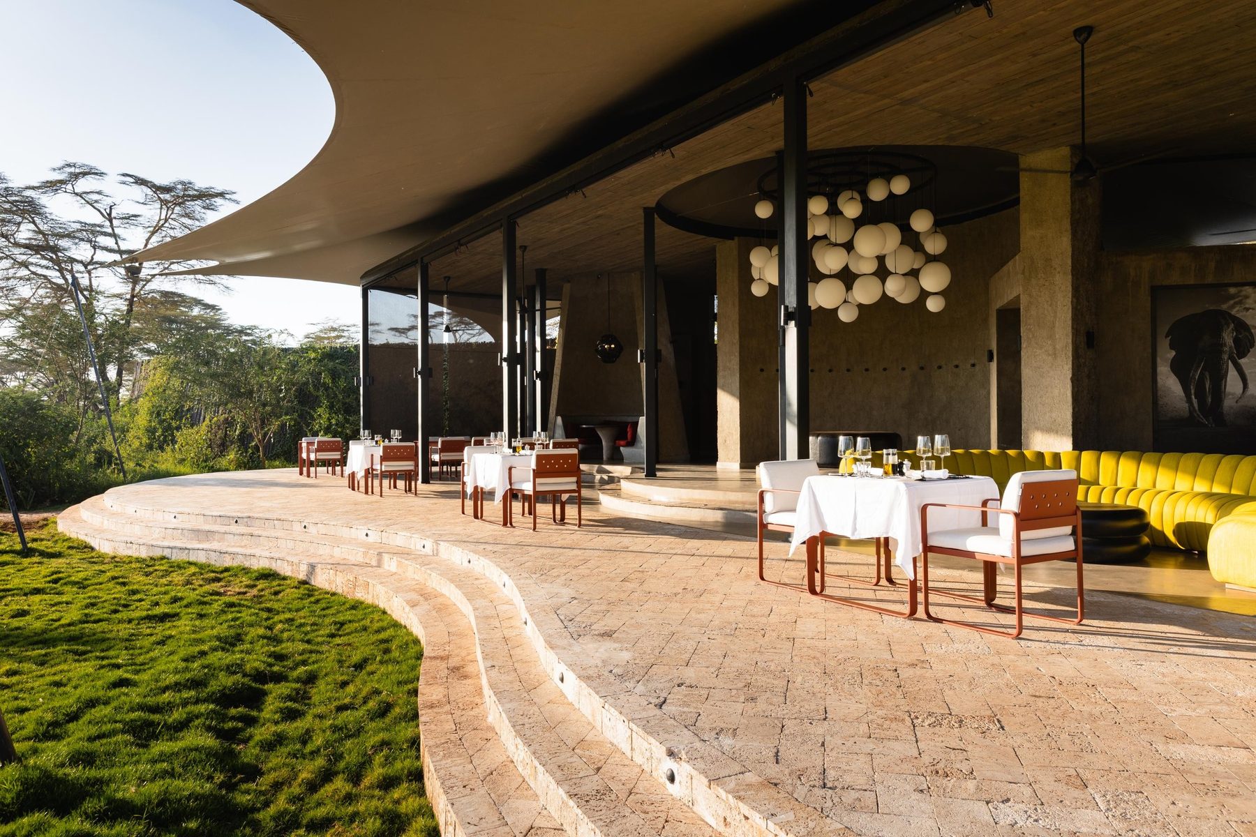 Curving stone terrace with set dining tables and yellow seating under a wide roof at Angama Amboseli