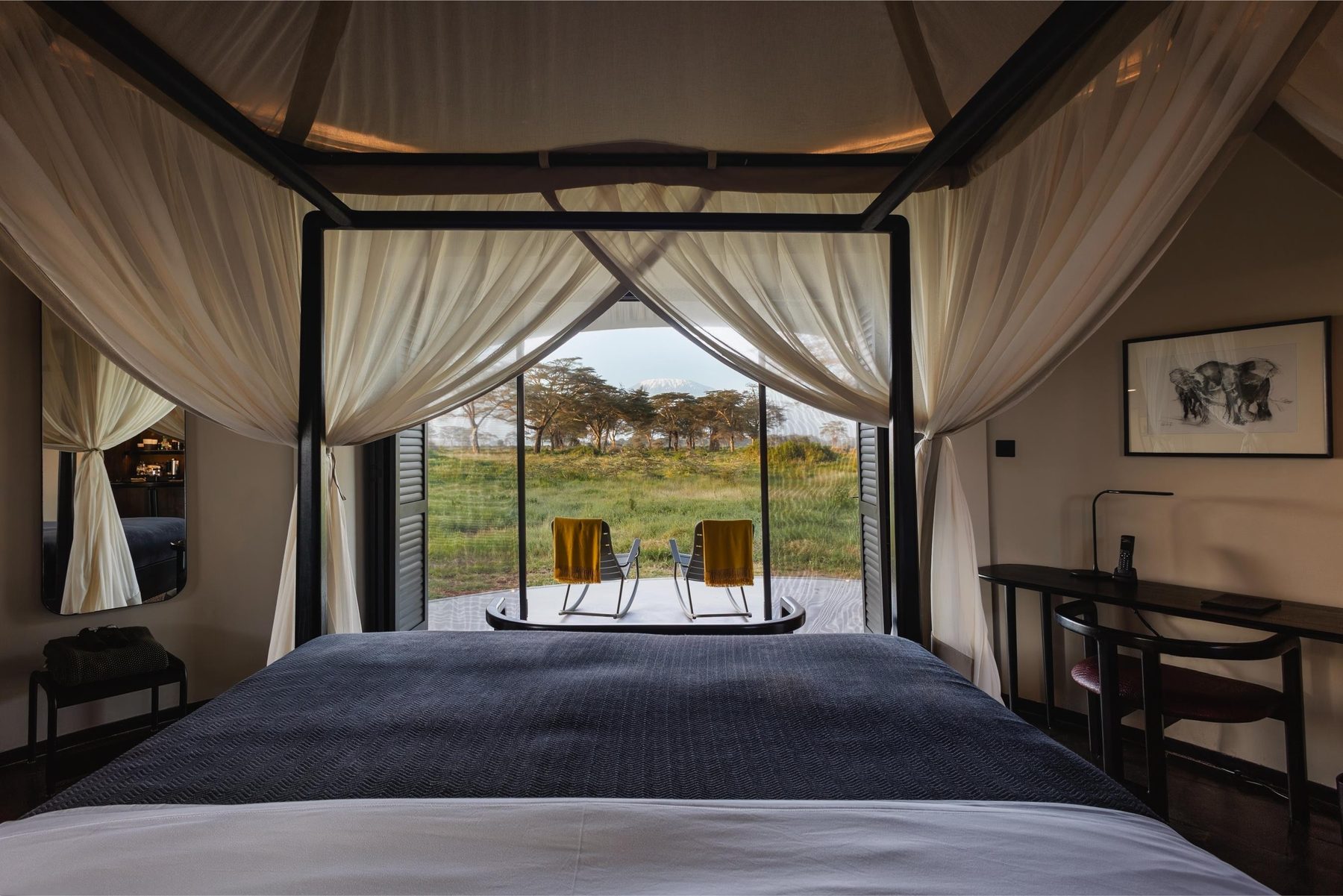 Bed under draped netting at Angama Amboseli, with curtains open to a deck with two chairs and green bush beyond
