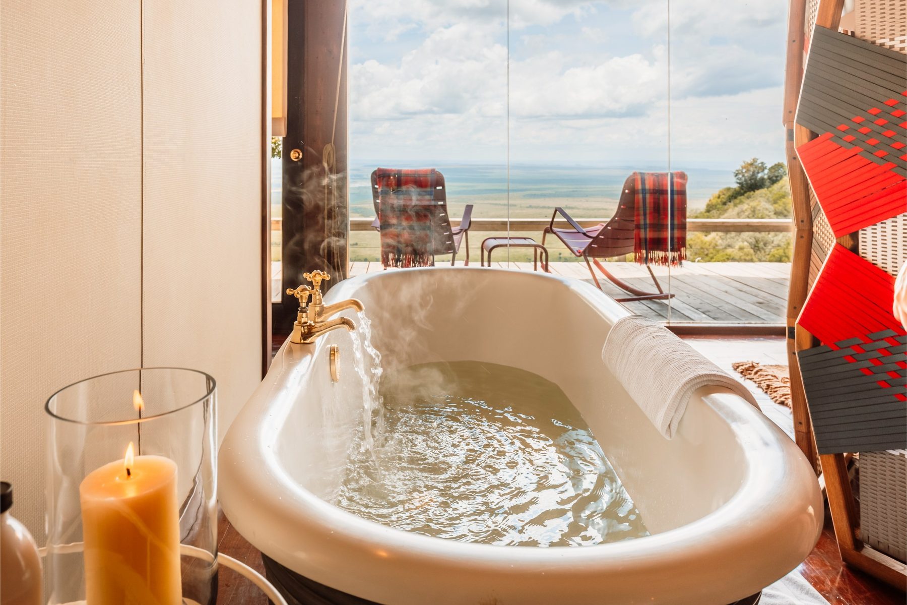 Freestanding bathtub with a lit candle beside glass doors and red deck chairs at Angama Mara