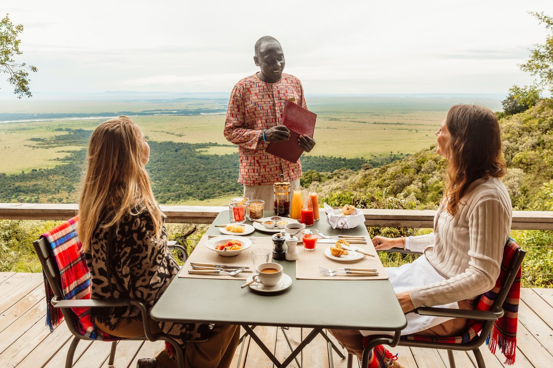 Two guests at breakfast on a deck high above the plains at Angama Mara, a host serving the table