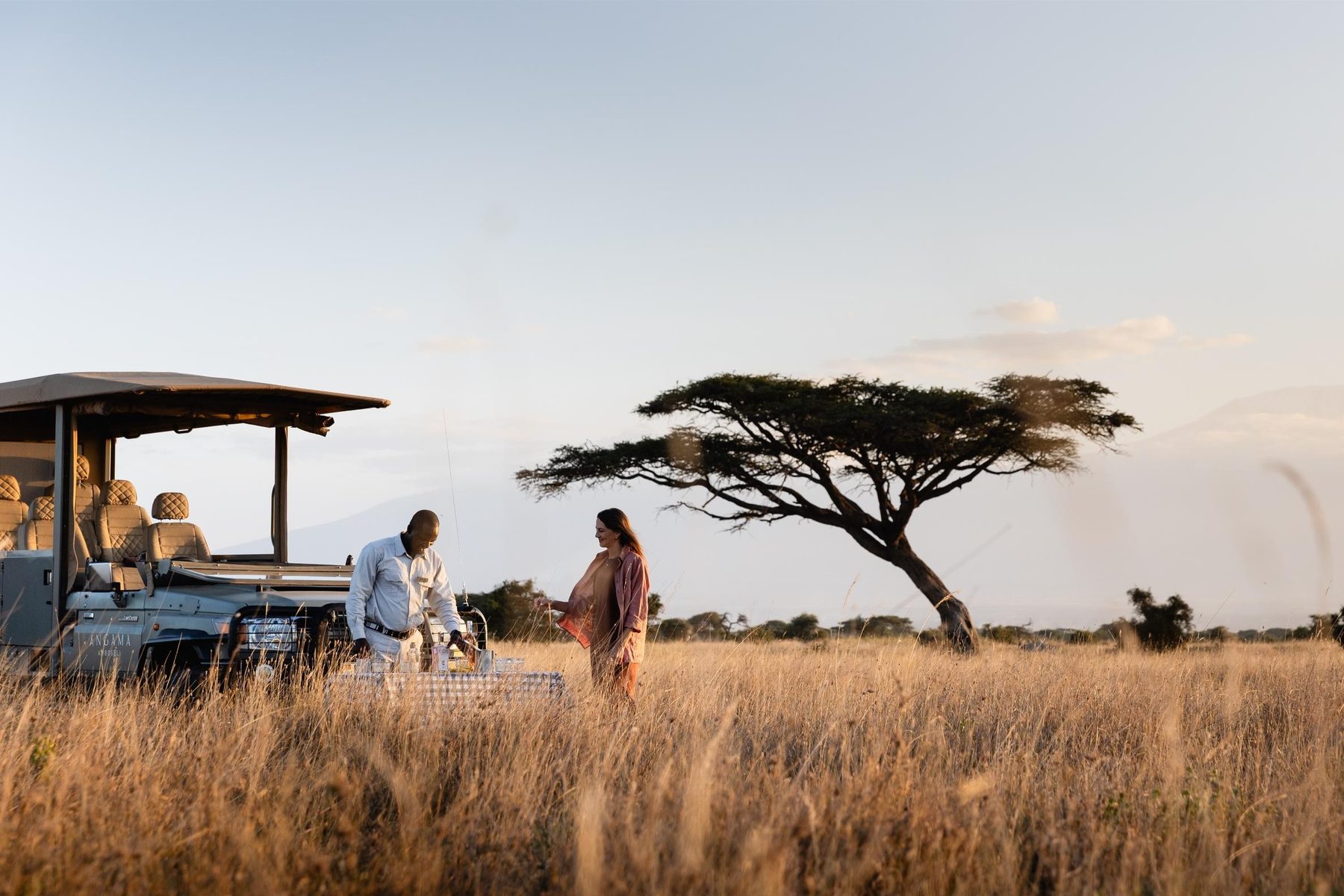 Guests stepping out of an open game drive vehicle into tall golden grass beside a lone acacia at Angama Mara