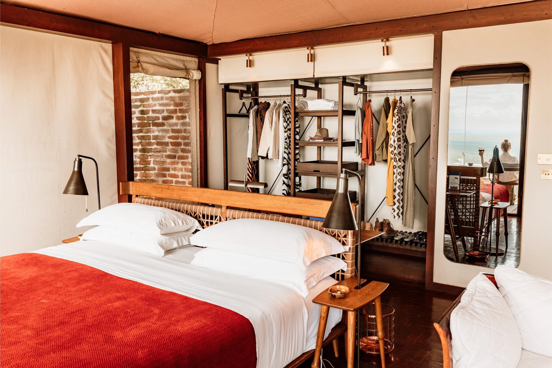 Bed with a red throw beside an open wardrobe and brick wall inside a tented suite at Angama Mara
