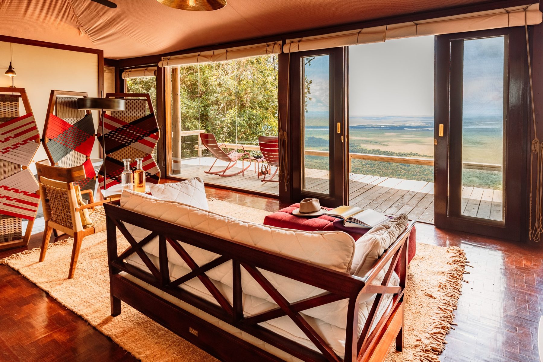 Suite at Angama Mara with a daybed, red ottoman and wooden floors, glass doors opening onto a deck above the plains