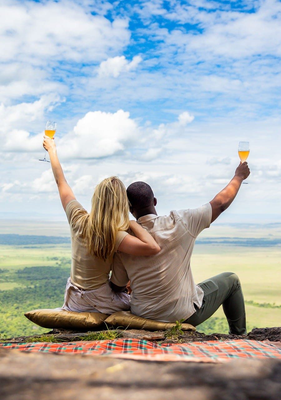 Couple raising glasses on a picnic blanket at the edge of an escarpment above the wide plains on an Angama outing
