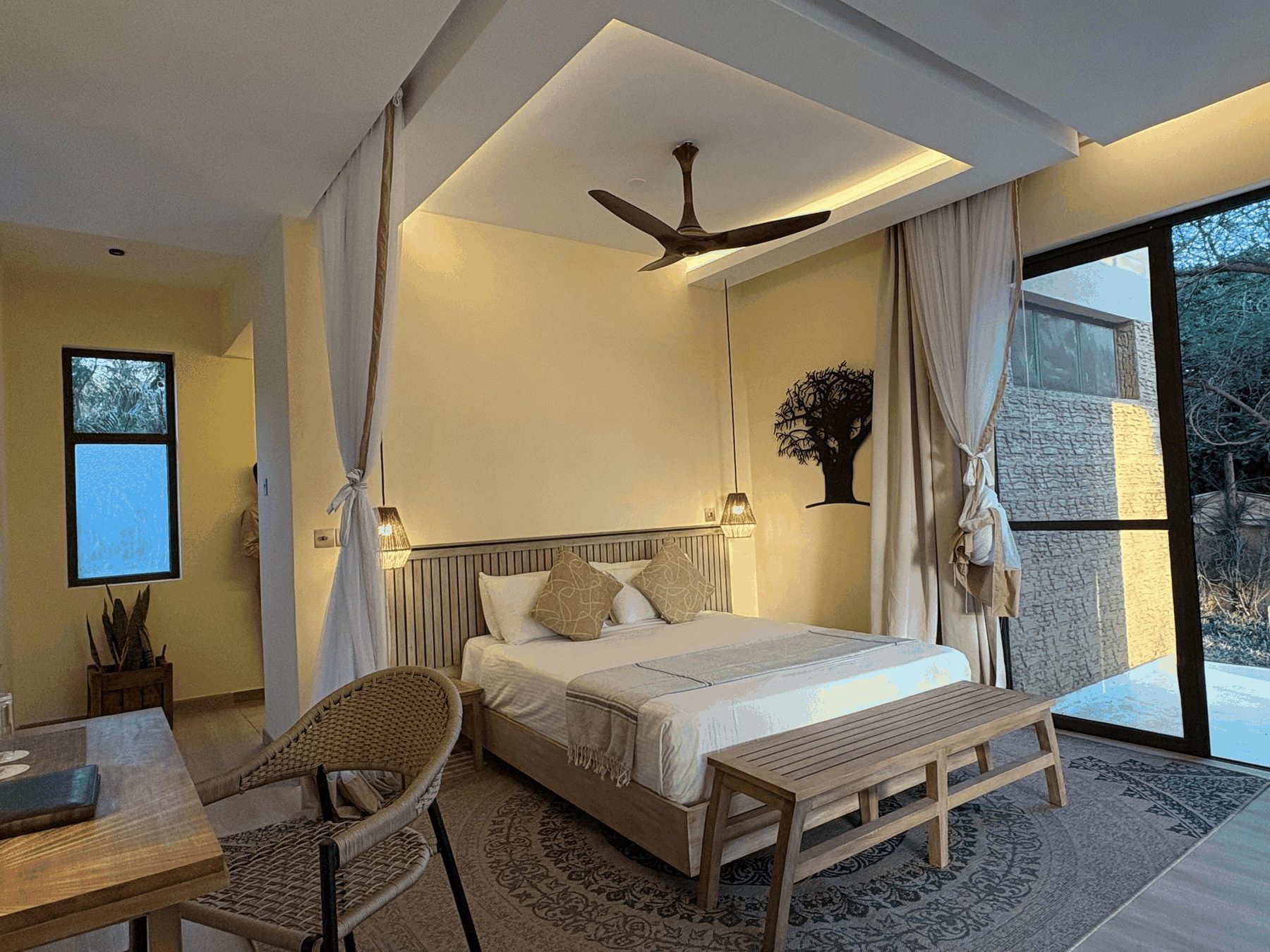 Bright bedroom at Ashnil Aruba Lodge with a ceiling fan, a woven rug, a wooden bench and a glass door to outside