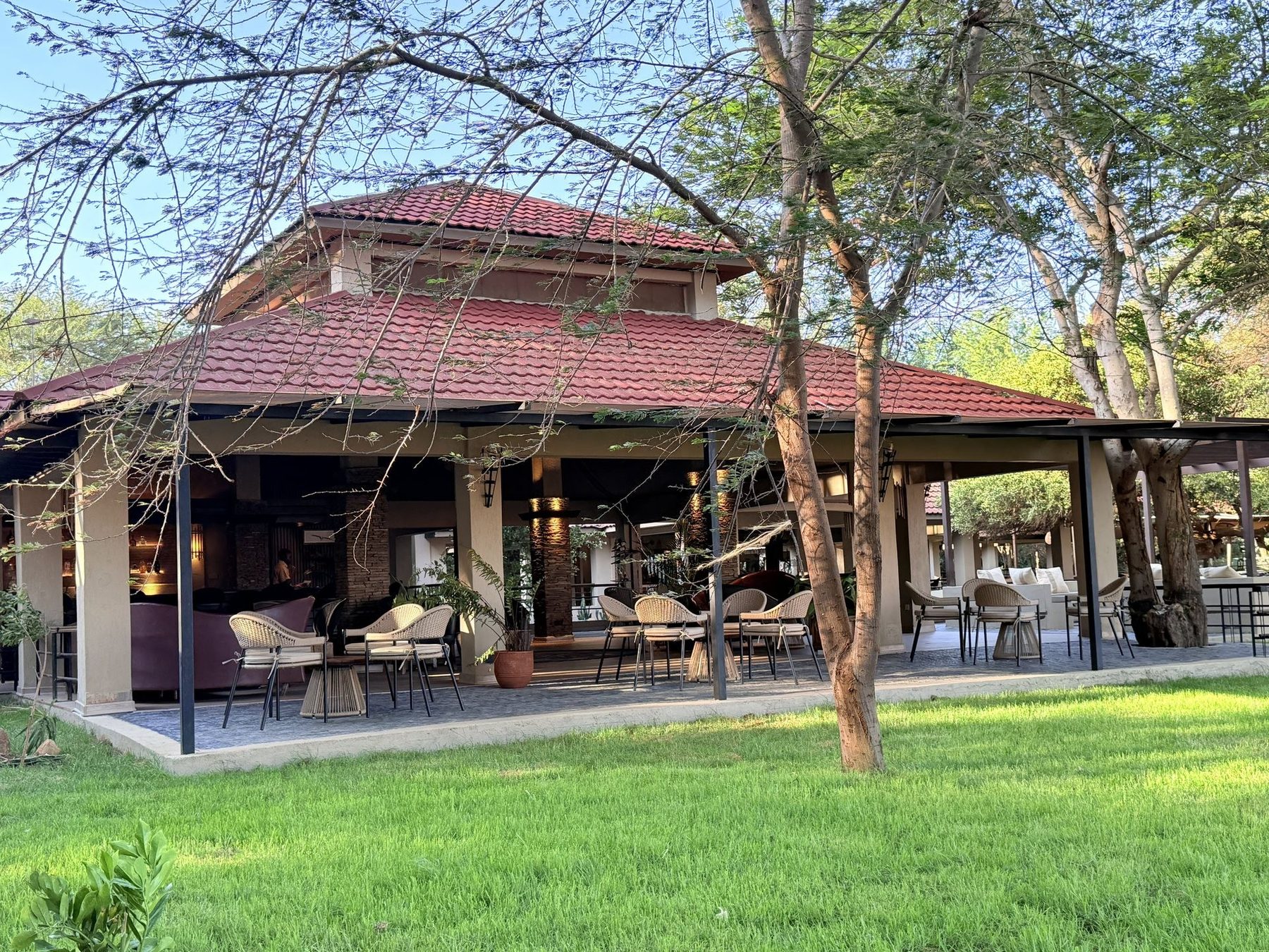 Open-sided main building with a red tiled roof at Ashnil Aruba Lodge, set on a green lawn among acacia trees
