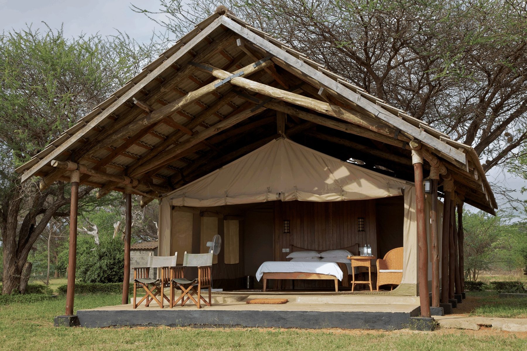 Canvas tent under a wooden thatch-style roof at Ashnil Aruba Lodge, with safari chairs on its veranda among trees