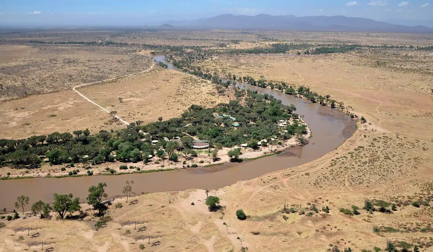 Aerial view of Ashnil Samburu Camp’s tents in a strip of trees along a winding river, with dry plains and hills beyond