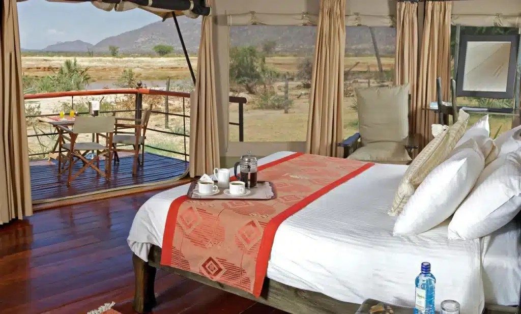 Tent at Ashnil Samburu Camp with a made-up bed, a tea tray and a wooden deck with chairs looking out to dry bush