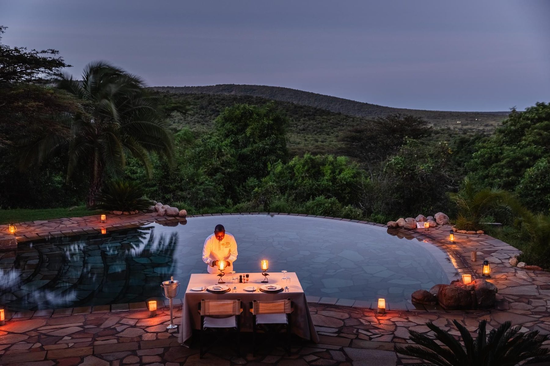 Candlelit dinner table set beside the pool at Cottar’s 1920s Camp at dusk, with wooded hills behind