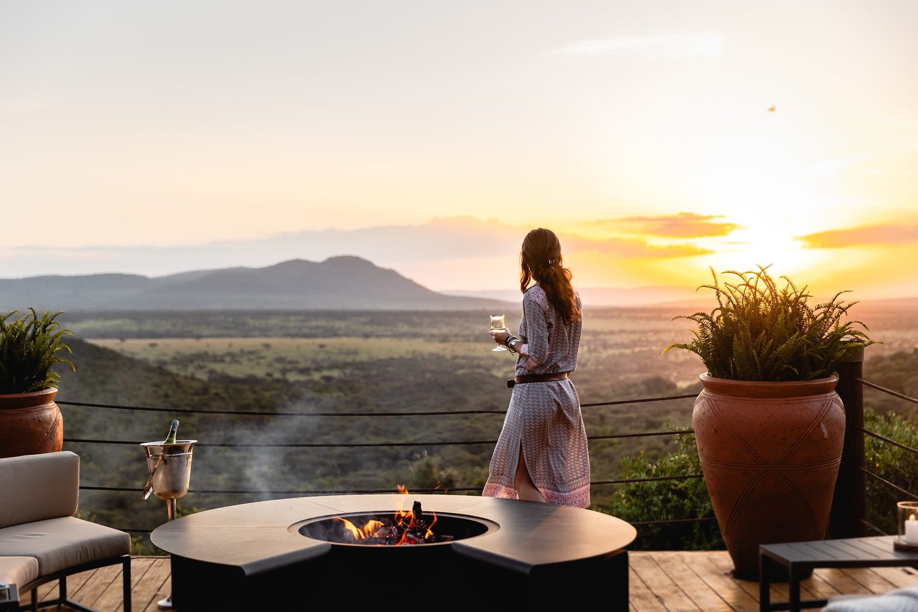 Guest with a glass by a fire pit on a deck at Cottar’s 1920s Camp, watching the sun set over the hills