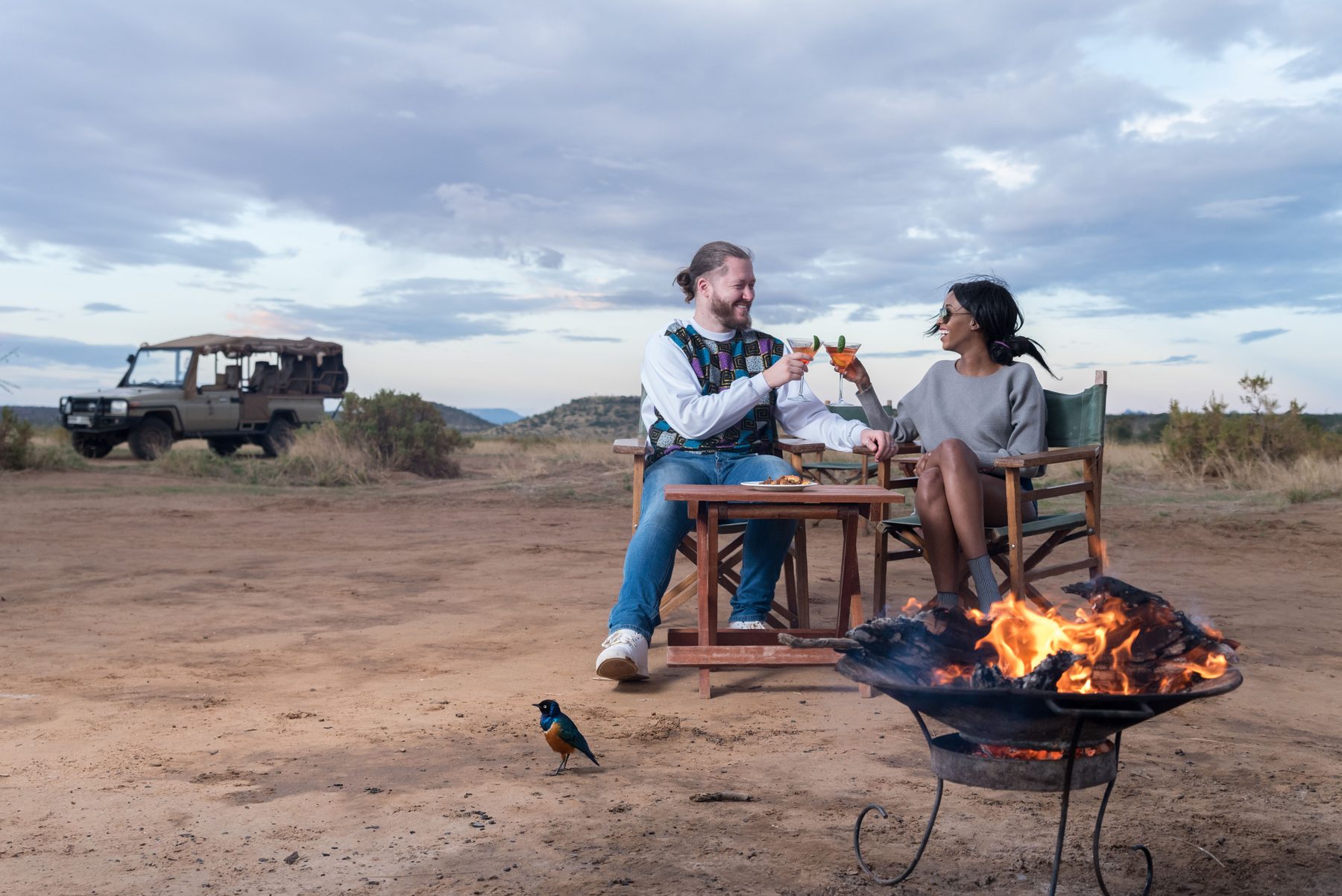 Couple toasting at a table beside a fire pit in open dry country, a game drive vehicle parked behind