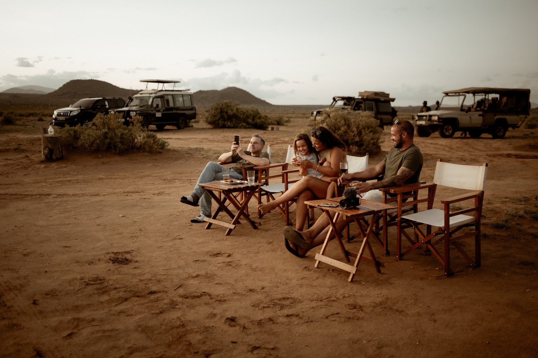 Guests relaxing in canvas chairs on red earth at dusk, with game drive vehicles parked behind them