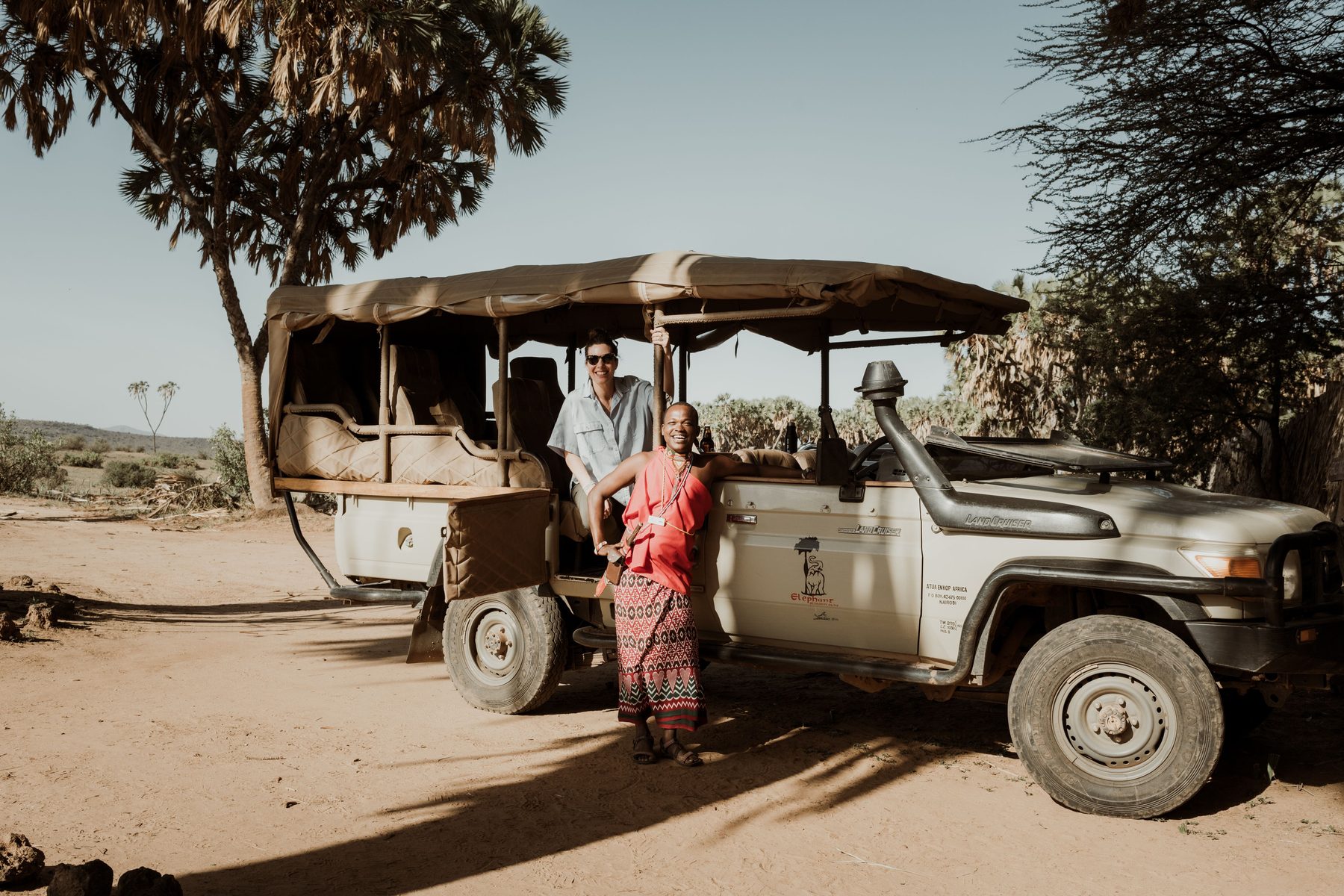 Guide in red shuka standing beside an open game drive vehicle in the shade of a tree at Elephant Bedroom Camp