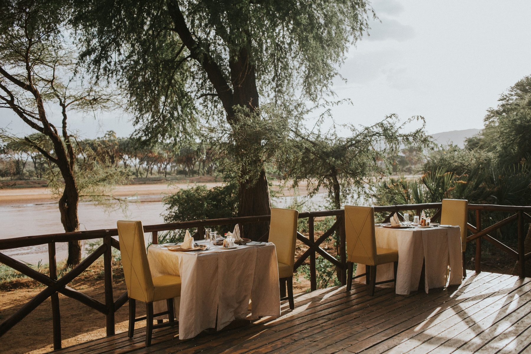Dining tables on a wooden deck under trees beside the Ewaso Nyiro River at Elephant Bedroom Camp