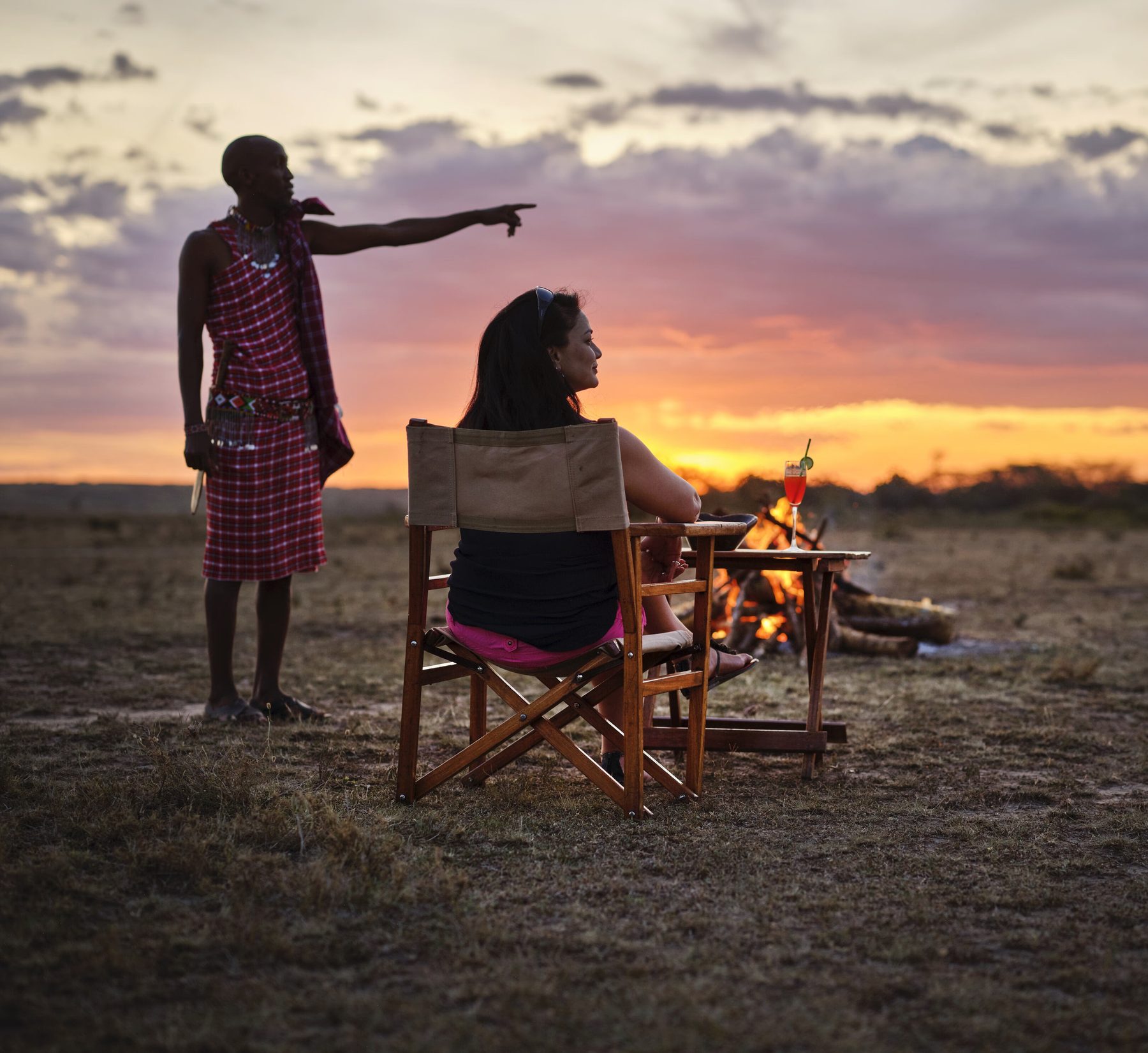 Guide pointing across the bush as a guest sits by a campfire under a glowing orange sunset