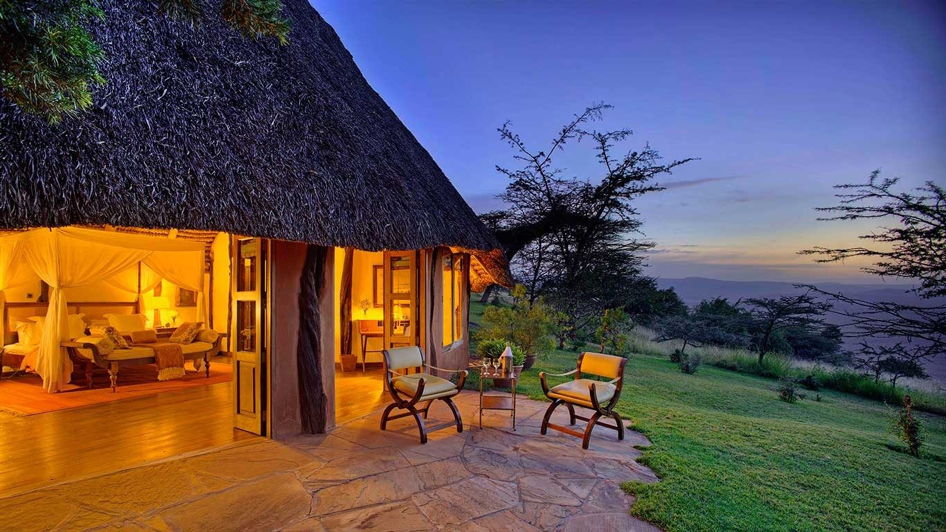 Thatched cottage at Elewana Kifaru House at dusk, lamps lit inside and chairs on a stone veranda above the lawn
