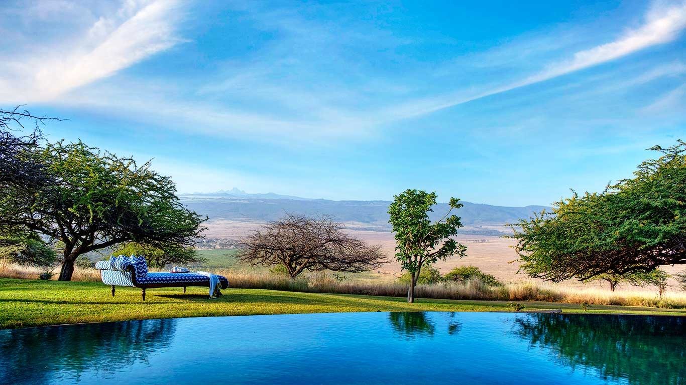 Still swimming pool reflecting acacia trees and blue sky at Elewana Kifaru House, with a lounger on the lawn