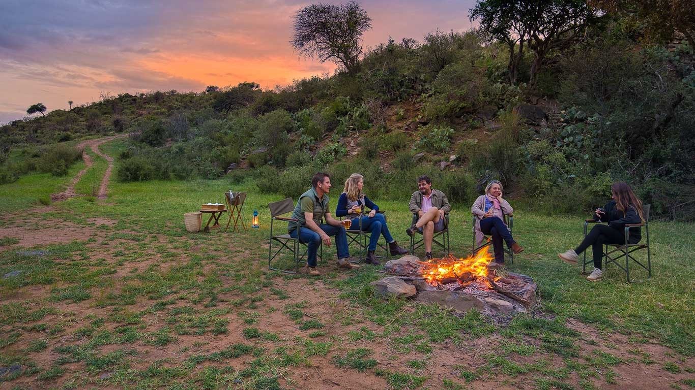 Guests sitting in camp chairs around a campfire on a grassy slope near Elewana Loisaba Tented Camp at dusk