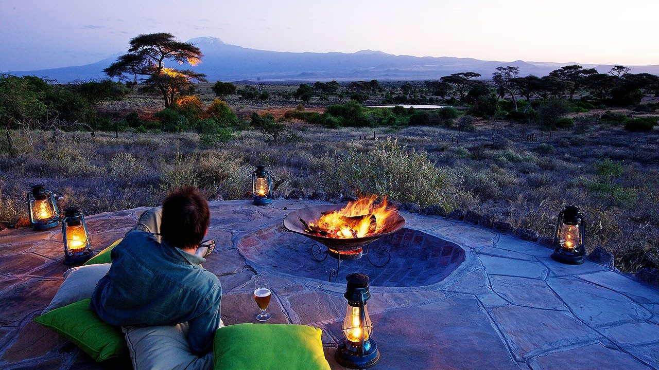 Guest relaxing on cushions beside a fire pit and lanterns on a stone terrace at Elewana Tortilis Camp Amboseli at dusk