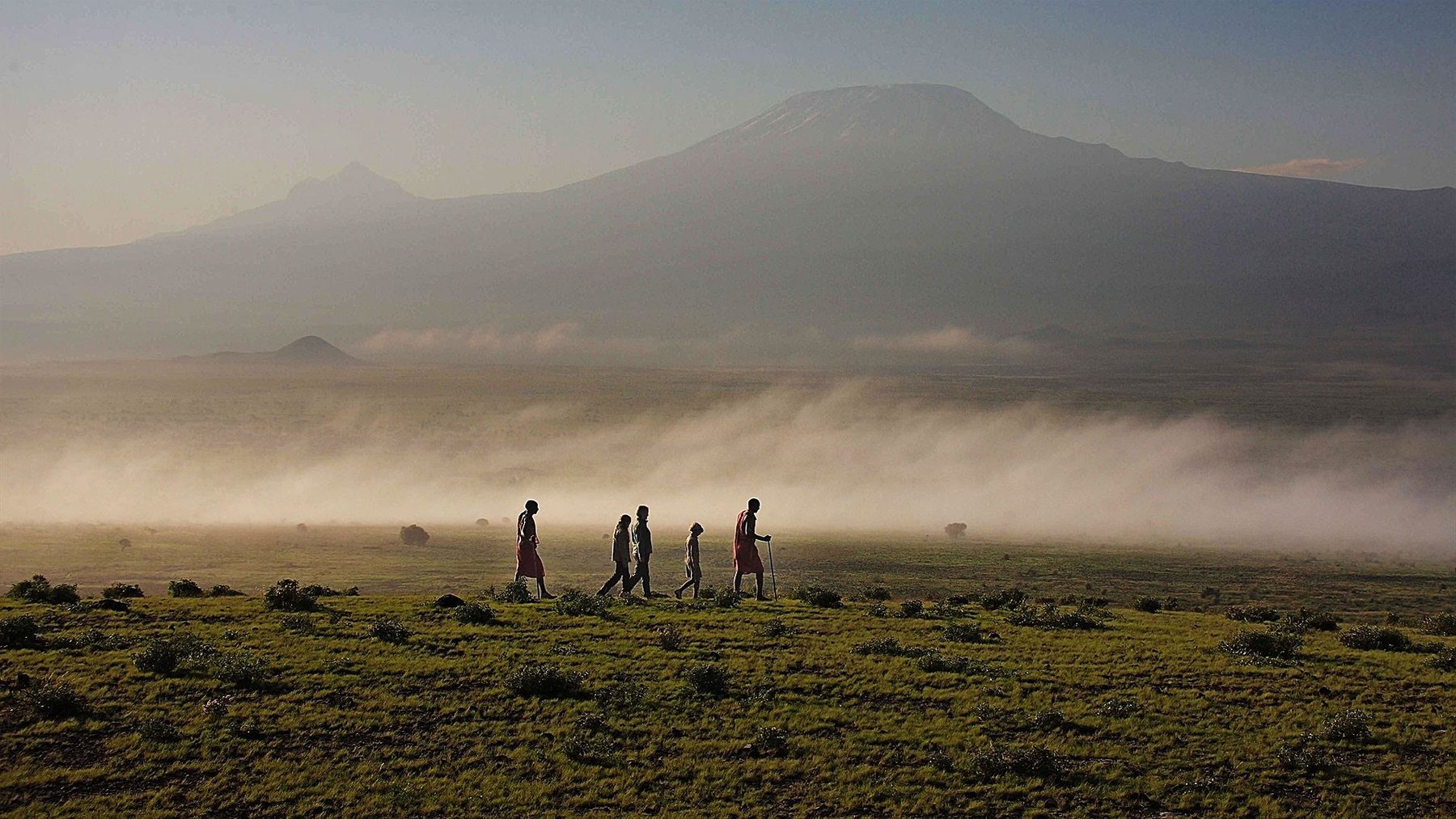 Small group walking across misty plains below the outline of Kilimanjaro near Elewana Tortilis Camp Amboseli