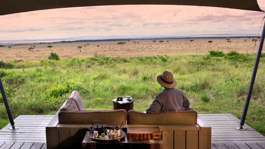 Guest in a hat on a deck at &Beyond Bateleur Camp, looking over plains dotted with trees and grazing herds