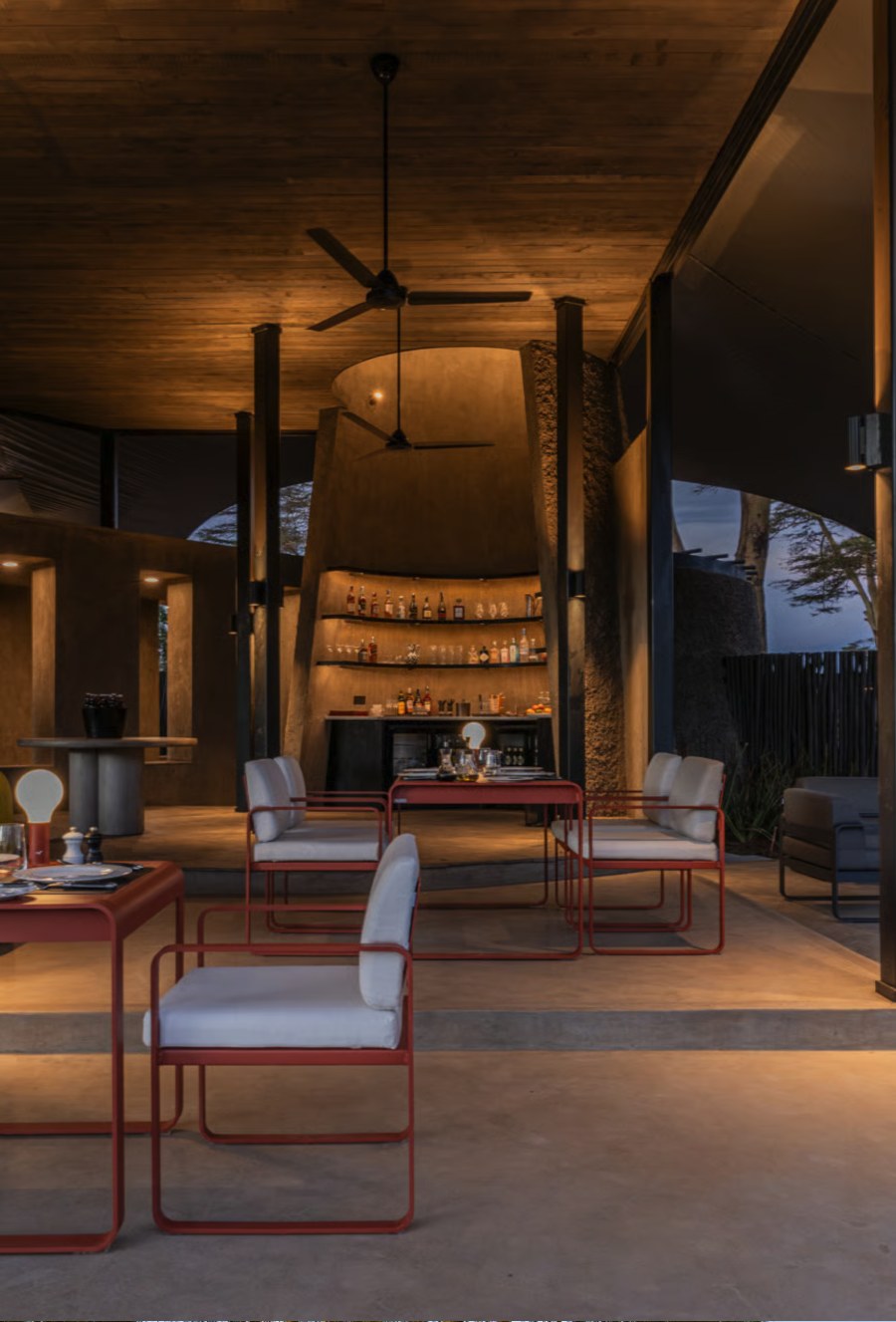 Dining area at Angama Amboseli at dusk with red-framed chairs, a stone bar and ceiling fans