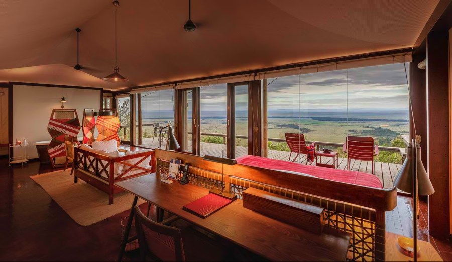 Glass-fronted tent at Angama Mara with a desk, sofa and a deck with red chairs looking over the plains