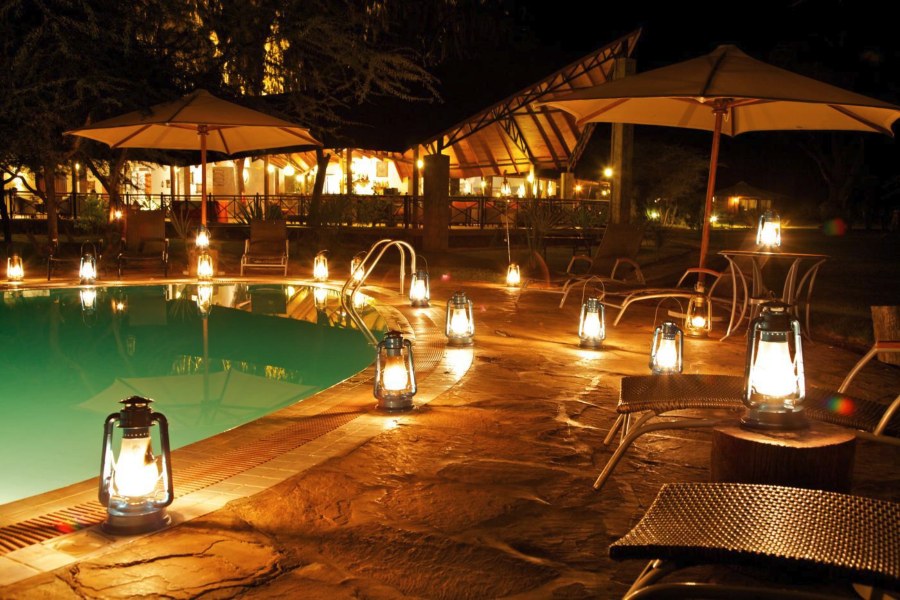 Pool at Ashnil Samburu Camp at night, lit by lanterns, with umbrellas and a thatched building behind
