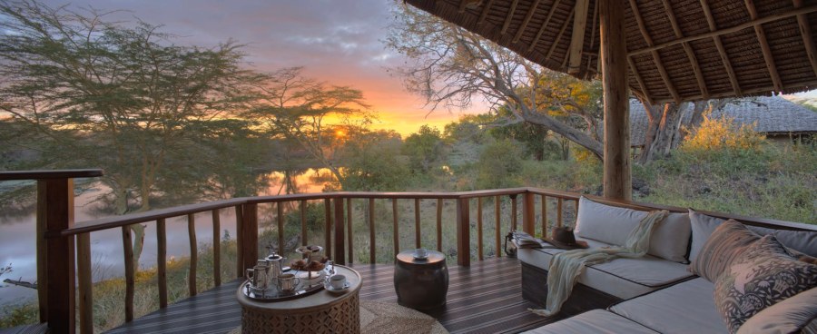 Thatched deck with cushioned seating and a tea tray at Finch Hattons, overlooking a pool among trees at sunset