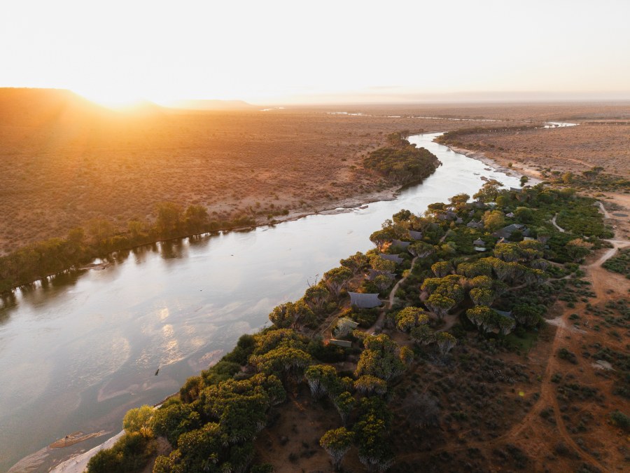 Aerial view of Galdessa Camp's roofs among green trees along the Galana River, with dry bush at sunrise
