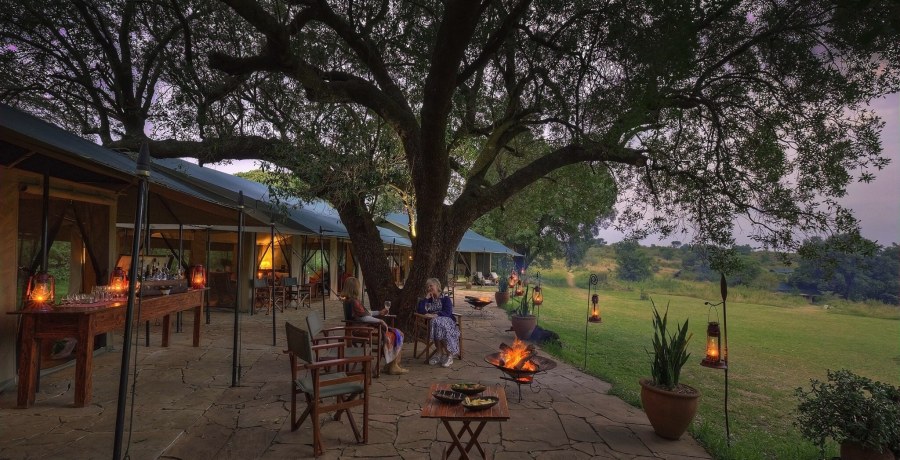 Guests by a fire pit on a stone terrace under a large tree at Kicheche Mara North, with lanterns and lawn