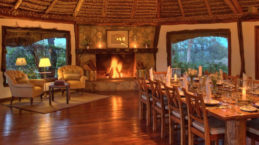Thatched dining room with a stone fireplace, armchairs and a long candlelit table at Elewana Kifaru House
