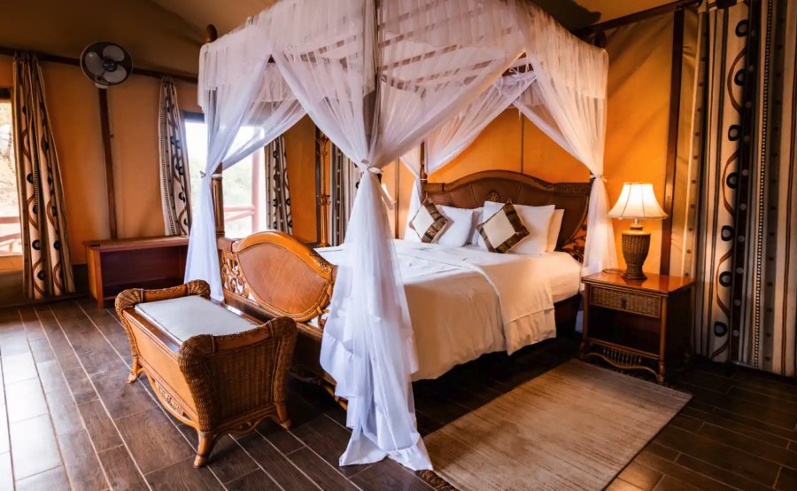 Tent at Kilima Safari Camp with a carved wooden bed under white netting, a bedside lamp and a cushioned bench