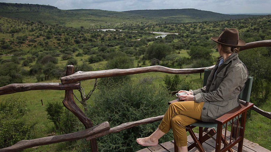 Guest with a cup of coffee on a wooden deck at Loisaba Star Beds, looking over green bush and a river valley