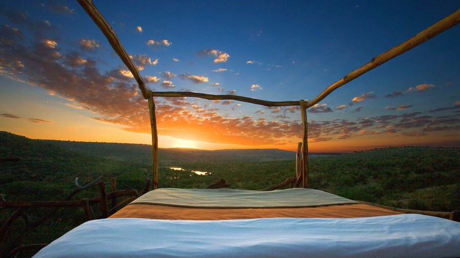 Open-air bed with a wooden frame at Loisaba Star Beds, facing the sunset over green bush and a river valley