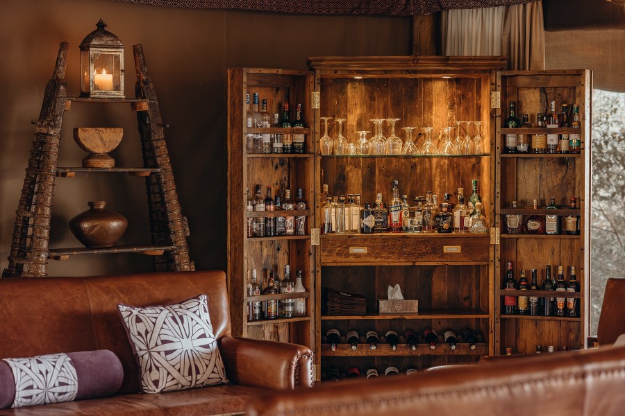 Wooden bar cabinet with bottles and glasses behind a leather sofa in the lounge tent at Mara Nyika Camp