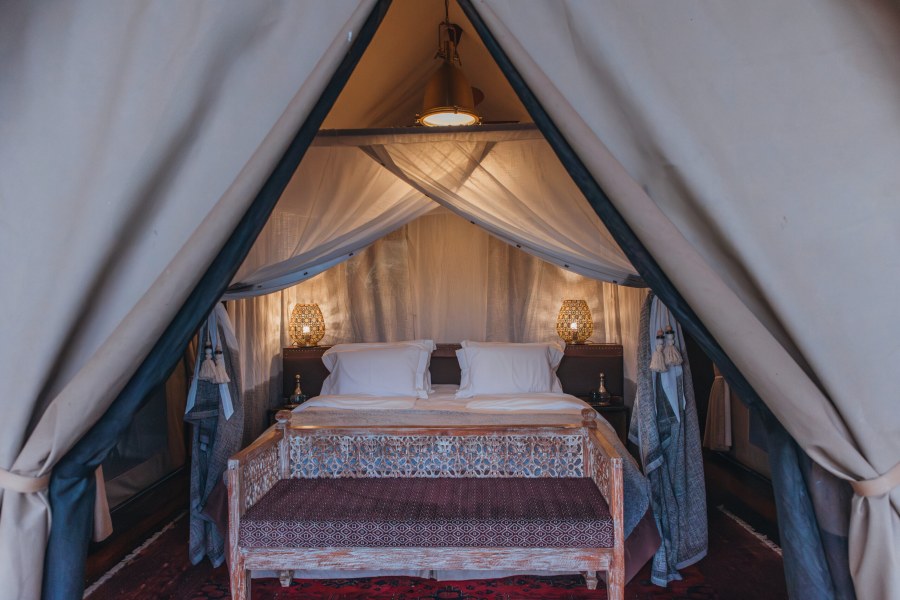 Tent flaps open onto a bed with white linen, carved bench and lamps under netting at Mara Nyika Camp