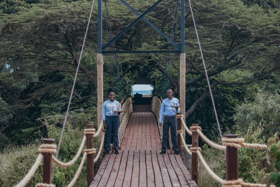 Two staff members holding trays on a rope suspension bridge among trees at Mara Plains Camp