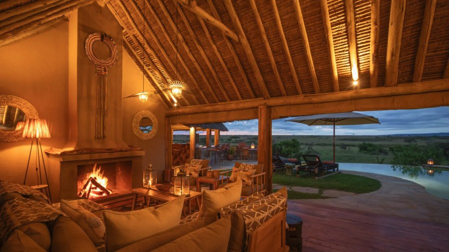 Thatched lounge with a lit fireplace and sofas at Naboisho Camp, opening onto a pool and the plains at dusk