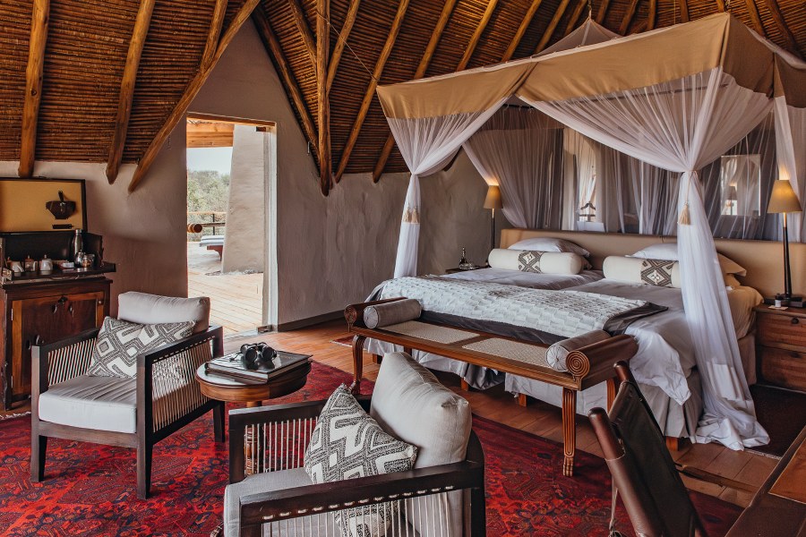 Thatched suite at ol Donyo Lodge with a canopy bed, armchairs on a red rug and a door open to a wooden deck
